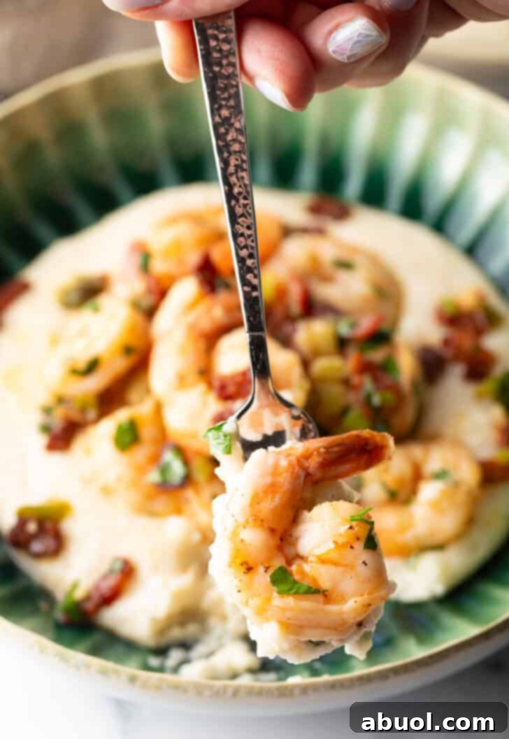 A green bowl brimming with cheesy grits and topped with Cajun shrimp and chopped bacon. A spoon holds a perfect bite of shrimp and grits, ready to be enjoyed, showcasing the dish's enticing flavors.