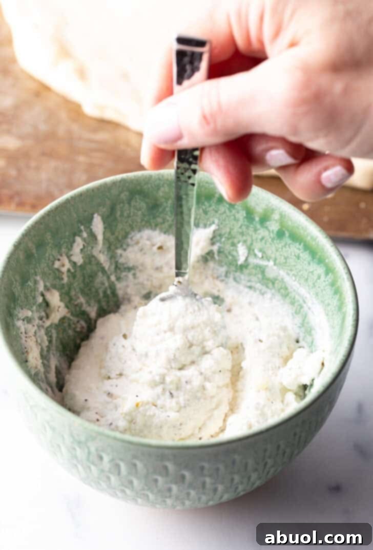Hand with a spoon stirring white sauce ingredients in a green bowl.