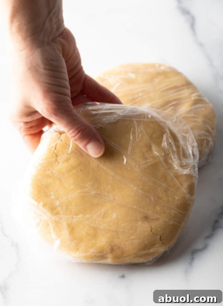 A hand holding a large, perfectly formed disc of pie dough, wrapped securely in plastic wrap for chilling.