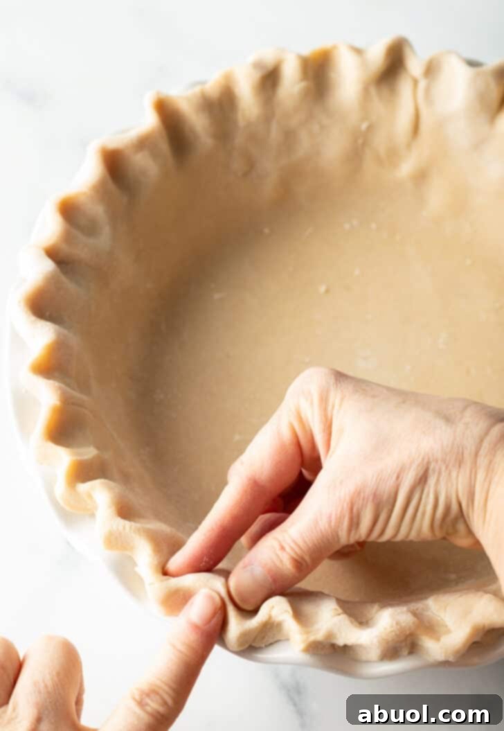 A hand meticulously crimping the raw pie dough around the edges of a pie plate, creating a decorative, sealed finish.