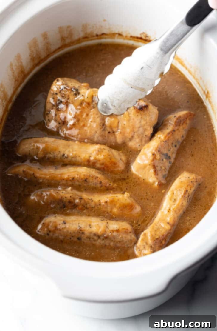 Tongs grabbing a pork chop from a white slow cooker filled with chops and onion gravy.