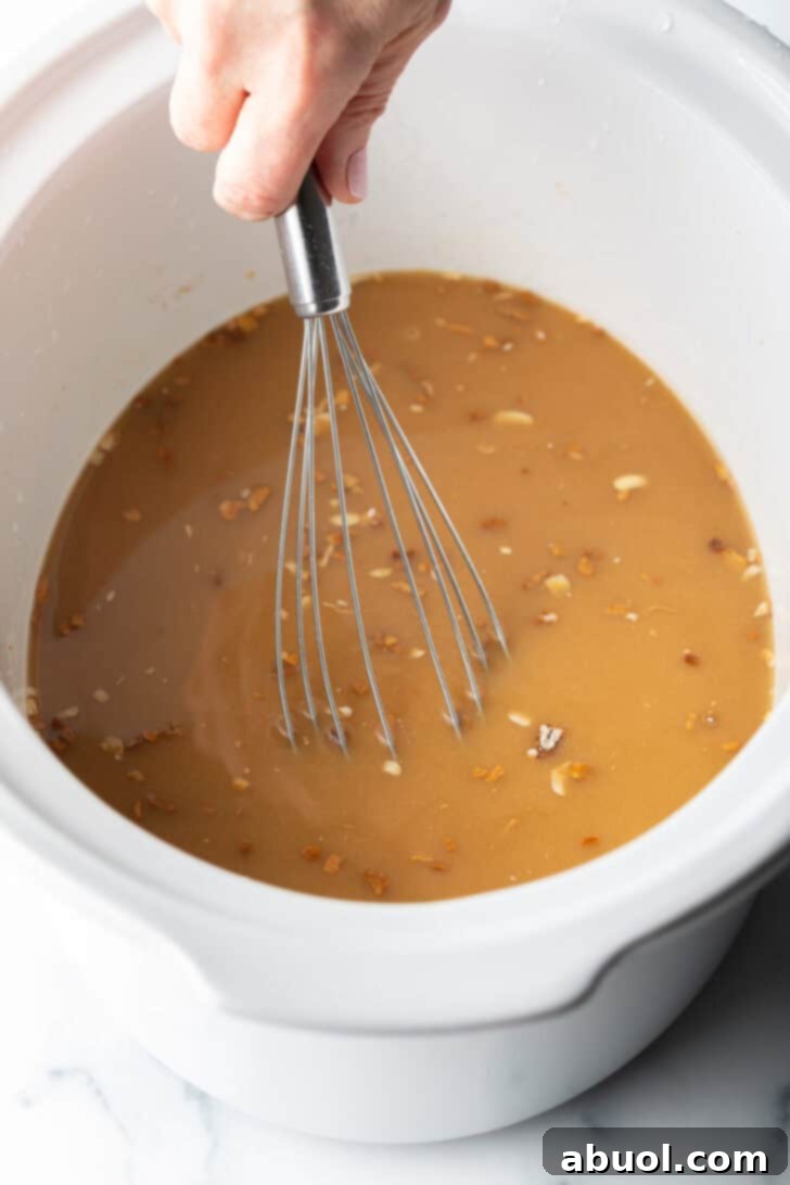Hand with whisk, whisking brown onion gravy in a white slow cooker.