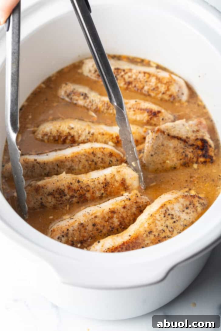 Tongs grabbing a pork chop from a white slow cooker filled with chops and onion gravy.