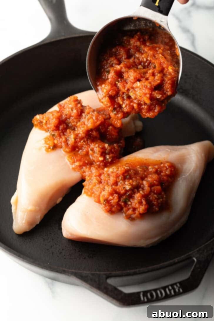 Adding tomato salsa from a measuring cup onto two raw chicken breasts in a cast iron skillet.