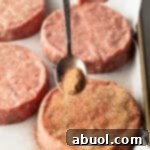 Sprinkling burger seasoning onto a raw hamburger patty with a metal spoon.