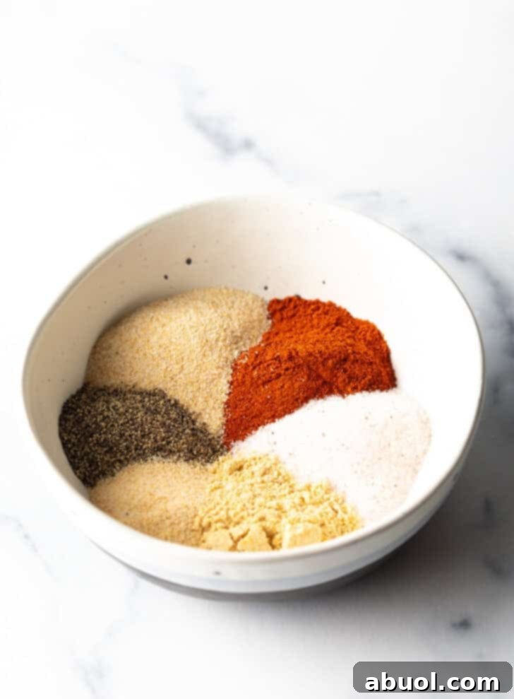 Individual piles of smoked paprika, salt, onion powder, black pepper, garlic powder, ground mustard, and a small heap of cayenne pepper artfully arranged in a white bowl, showcasing the fresh spices.
