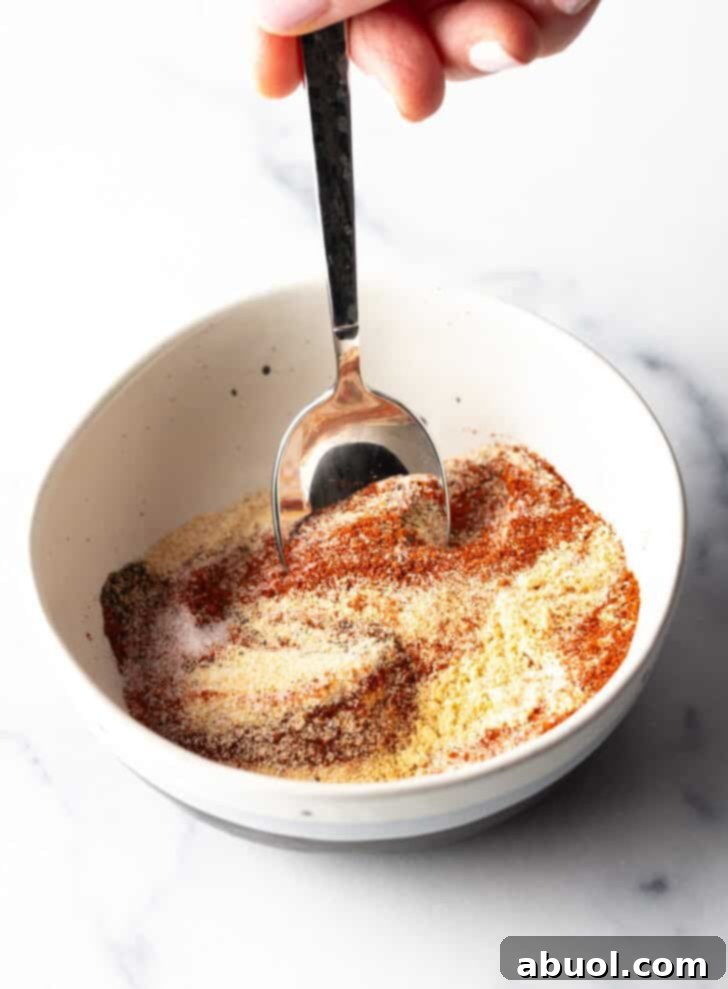 A hand stirring together a mixture of spices in a white bowl with a spoon, creating the homemade burger seasoning.