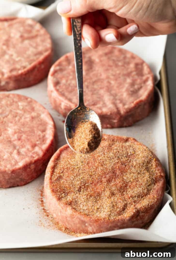 A metal spoon delicately sprinkling a generous amount of burger seasoning onto a raw hamburger patty, ensuring even coverage.