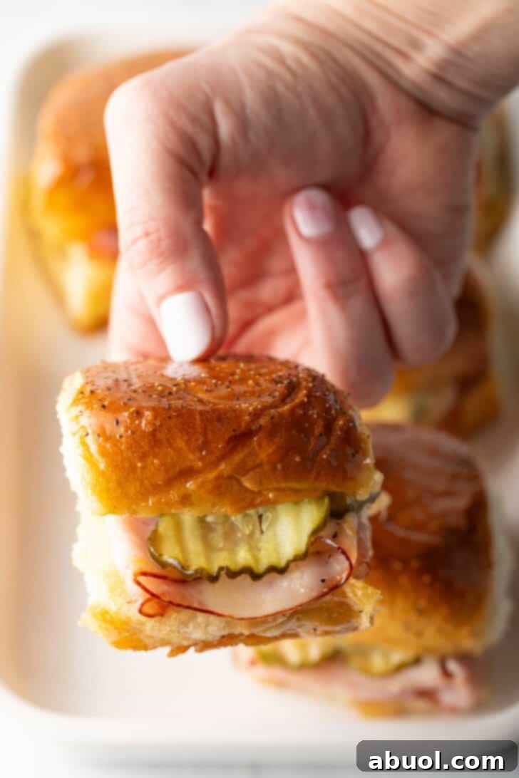 Sweet Hawaiian Bites 13 A hand gently holding up a single King's Hawaiian ham and cheese slider, showing off the layers of ham, melted cheese, and a crisp pickle slice within.