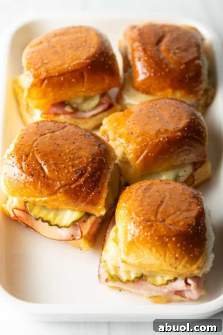 Sweet Hawaiian Bites 4 Five King's Hawaiian ham and cheese sliders neatly arranged on a white rectangular plate, showcasing their golden-brown tops and slightly melted cheese.