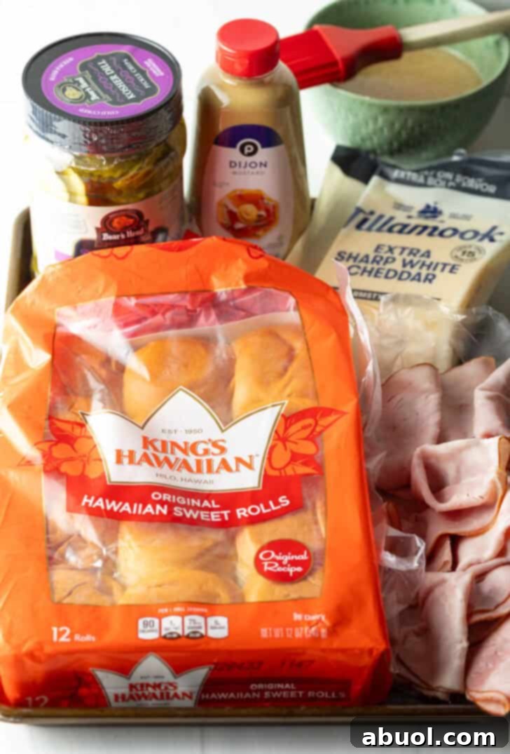 Sweet Hawaiian Bites 5 A vibrant flat lay of King's Hawaiian slider ingredients: a bag of sweet rolls, a stack of deli ham, slices of white cheddar cheese, a bottle of Dijon mustard, and a jar of dill pickles.