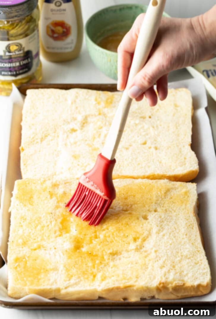 Sweet Hawaiian Bites 6 A hand using a red silicone brush to generously coat the bottom layer of King's Hawaiian rolls with melted honey butter.