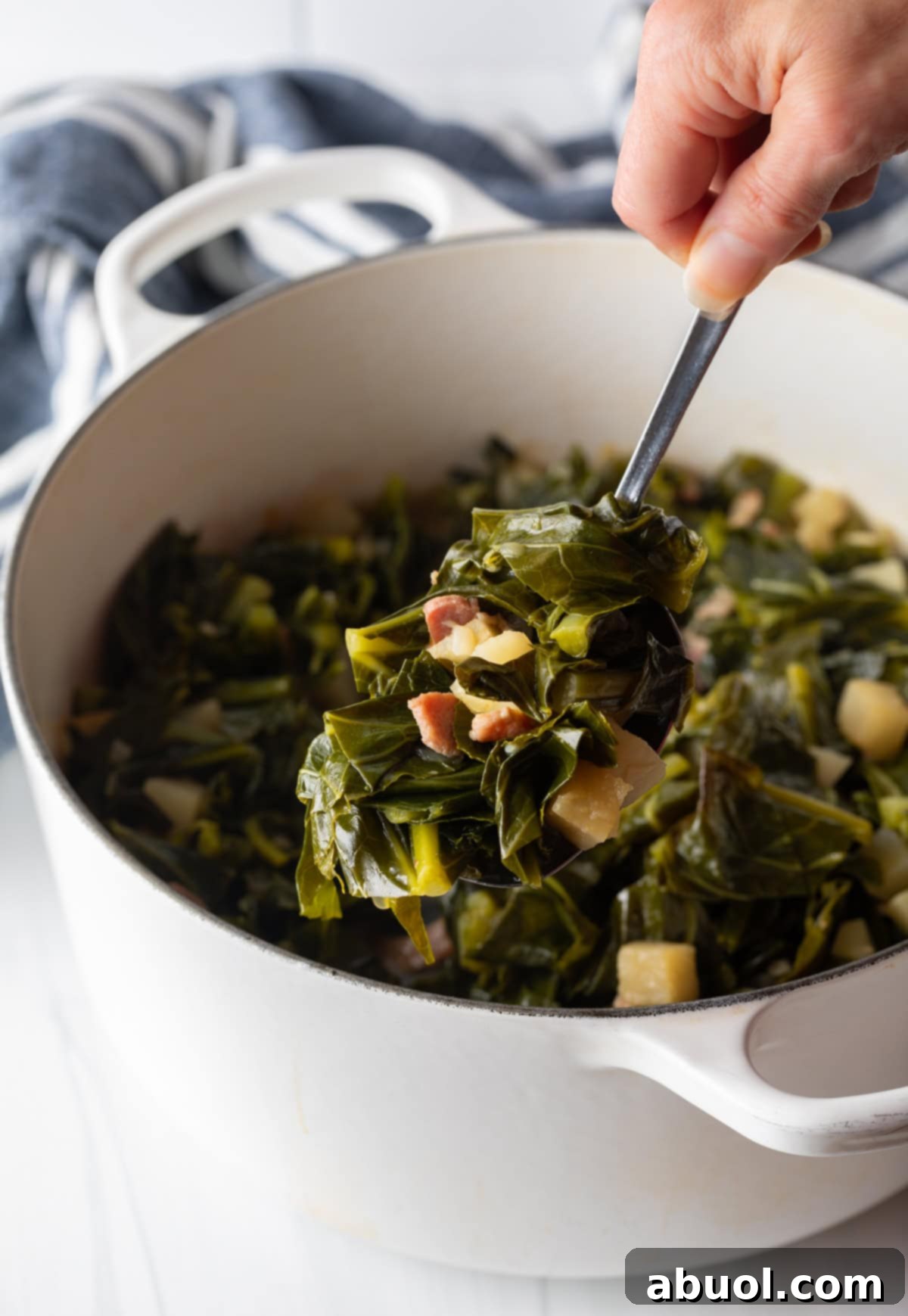 A close-up shot of a hand holding a spoonful of tender collards, with a large pot of the rich greens visible in the softly blurred background.