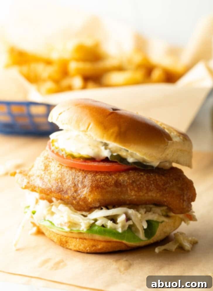 Battered and fried fish sandwich on a bun with coleslaw, tartar sauce, and a slice of tomato.
