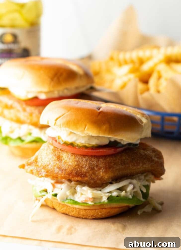 Two battered and fried fish sandwiches on a bun with coleslaw, tartar sauce, and a slice of tomato.