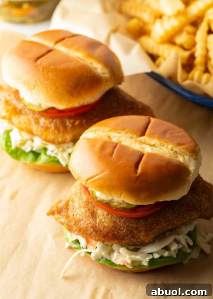 Two battered and fried fish sandwiches on a bun with coleslaw, tartar sauce, and a slice of tomato.