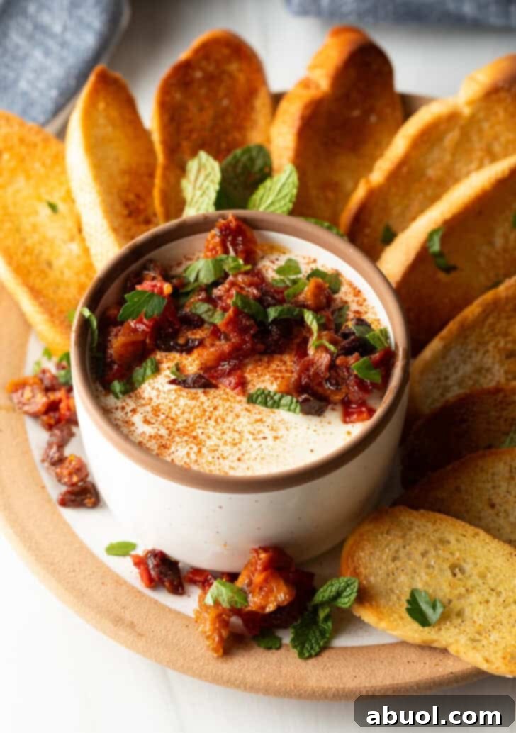 White cup with whipped feta with chopped sun dried tomato, on a plate surrounded by slices of toasted baguette.