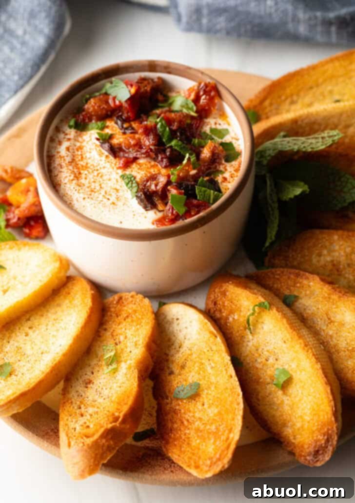 White cup with whipped feta with chopped sun dried tomato, on a plate surrounded by slices of toasted baguette.