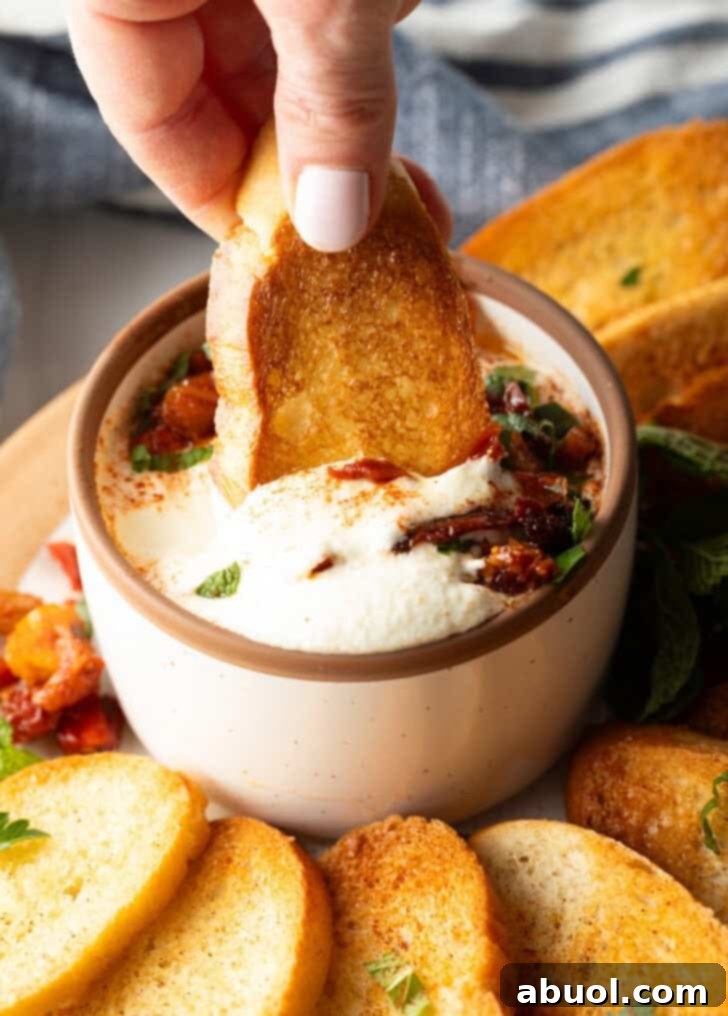 Hand dipping a slice of toasted baguette into a bowl of creamy whipped feta dip.