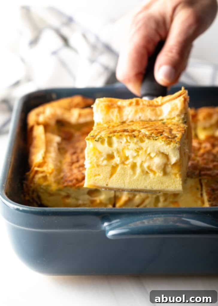 A hand is carefully removing a perfectly baked square of egg casserole from a square baking dish using a spatula, revealing its golden-brown top and fluffy interior.