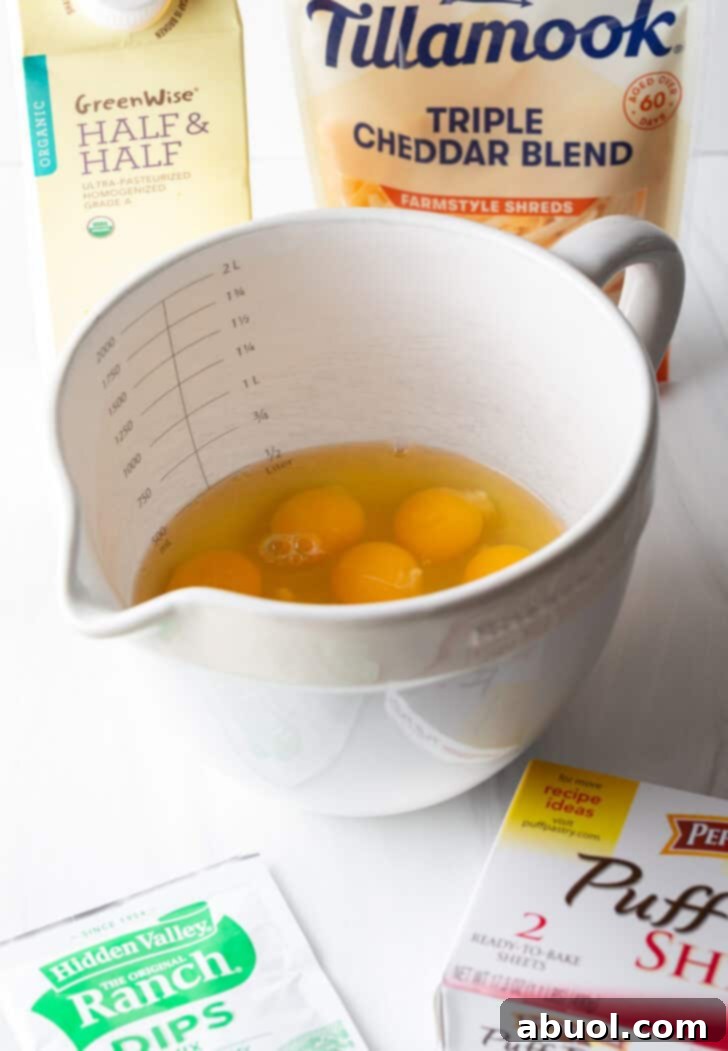 All the essential ingredients for the easy breakfast casserole are neatly arranged: a large white mixing pitcher filled with eggs, a box of frozen puff pastry, a packet of ranch dressing mix, a bag of shredded cheddar cheese, and a carton of half and half.