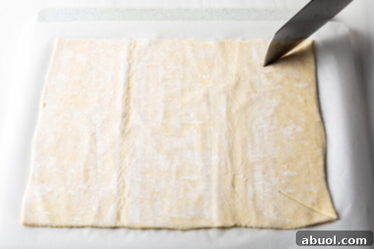 A sharp knife precisely slices a large rectangle of thawed puff pastry dough on a flat surface.