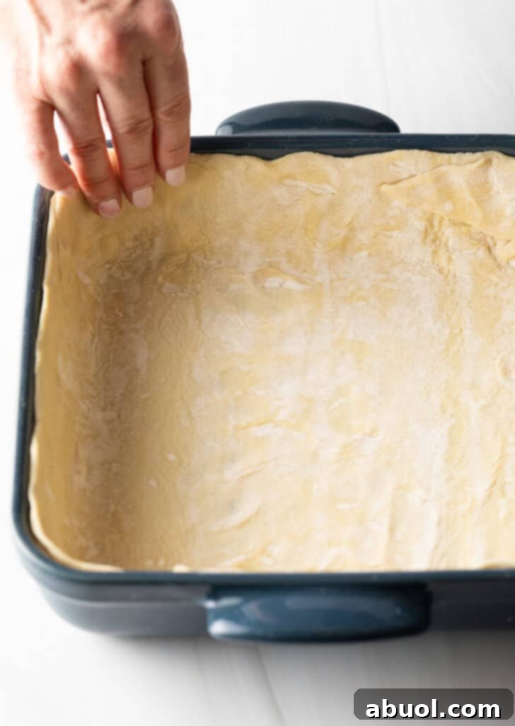 A hand gently presses the puff pastry dough into the bottom and up the sides of a square baking dish, forming the crust.