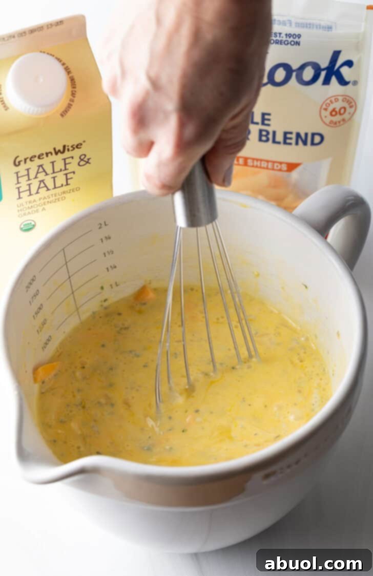 A hand vigorously whisks eggs and shredded cheese together in a large white mixing pitcher, creating a uniform mixture.
