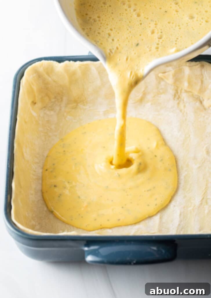 The blended egg mixture is steadily poured from a measuring cup into a prepared baking dish, which is already lined with a golden puff pastry crust.