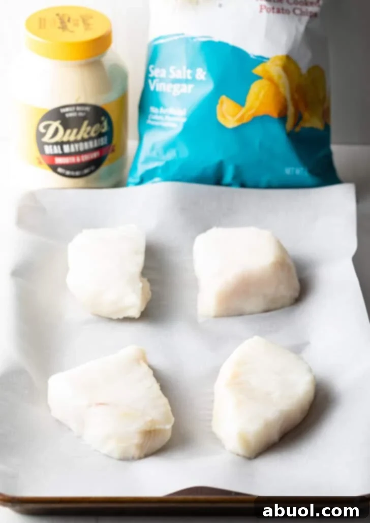 Four pristine raw sea bass fillets neatly arranged on white parchment paper, with a vibrant bag of salt and vinegar potato chips and a jar of creamy mayonnaise blurred in the background, highlighting the simple yet powerful ingredients.