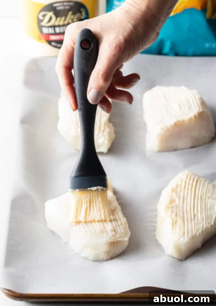 A black basting brush evenly coating four fresh white grouper fillets with a thin layer of mayonnaise on a baking sheet lined with parchment paper, preparing them for the crispy crust.