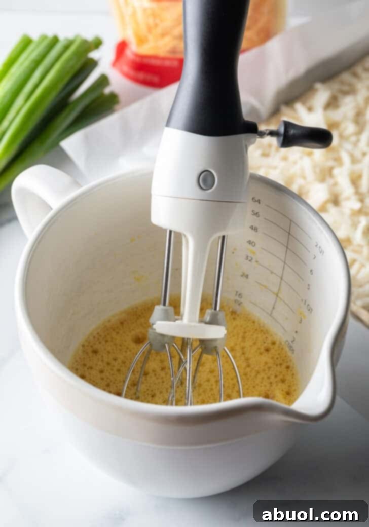 A white mixing pitcher containing a frothy egg mixture, with a hand holding a whisk, illustrating the egg preparation step.