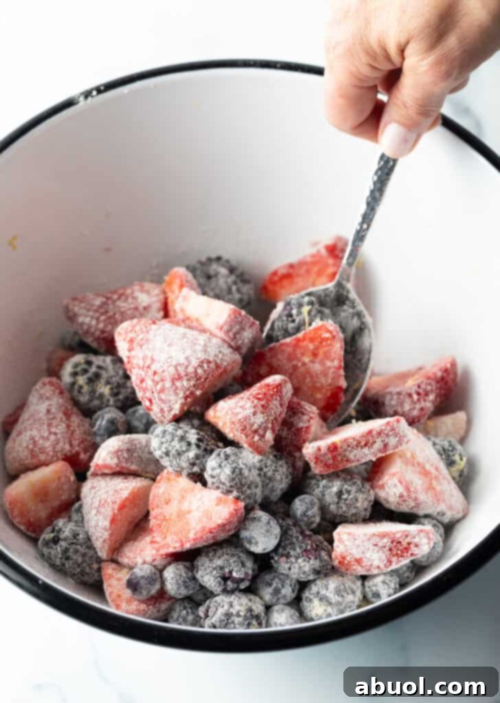Tossing a colorful mix of strawberries, blueberries, and blackberries with sugar, flour, and lemon zest in a white bowl.