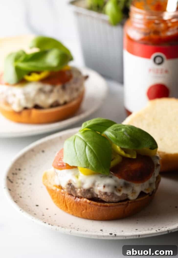 A complete open-face pizza burger on a white and black speckled plate, beautifully adorned with the bottom bun, sauce, a burger patty with melted cheese and pepperoni, all topped with vibrant banana pepper rings and fresh baby basil leaves, ready to be enjoyed.