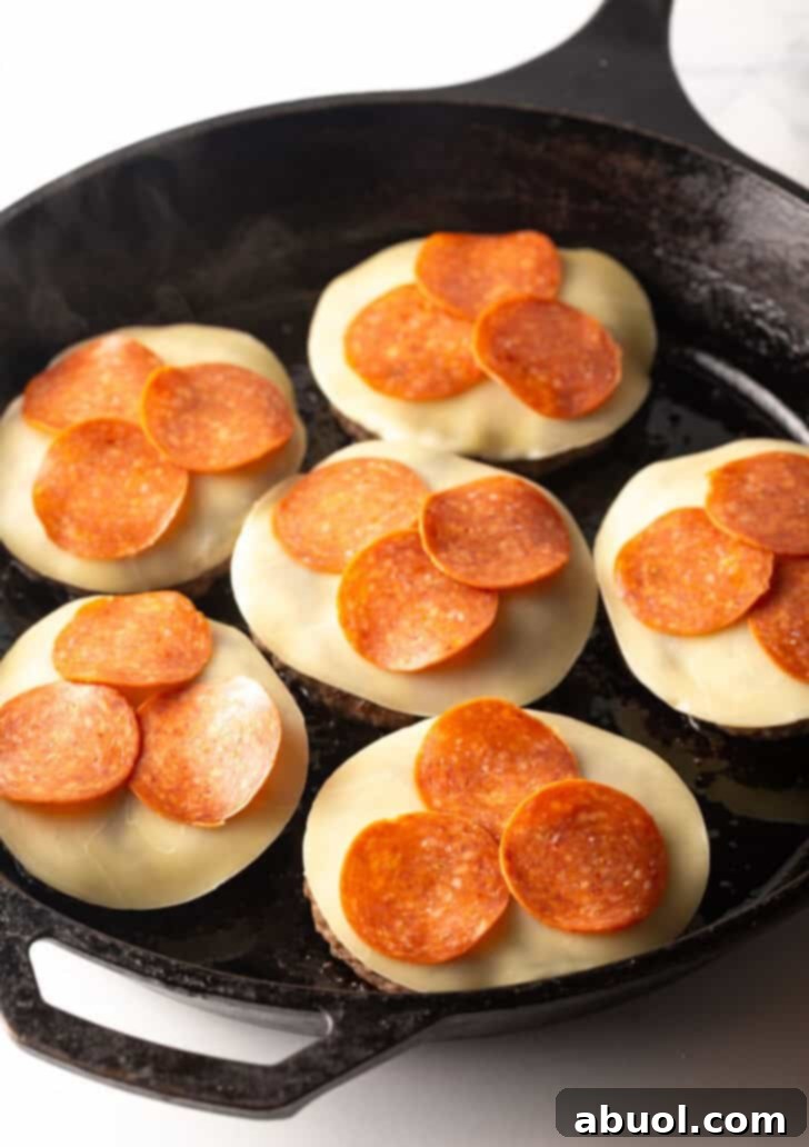 Six burger patties nestled in a cast-iron skillet, each generously topped with slices of mozzarella cheese and pepperoni, poised for melting.