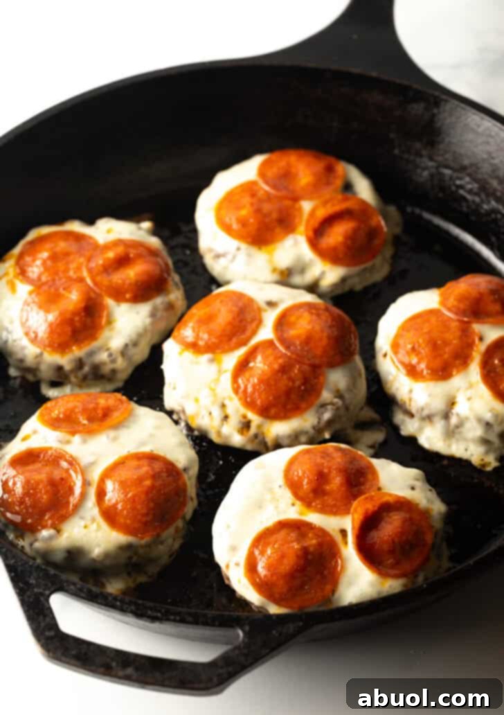 Six burger patties in a cast-iron skillet, now featuring perfectly melted mozzarella cheese and slightly crisped pepperoni slices, fresh and bubbly from the broiler.