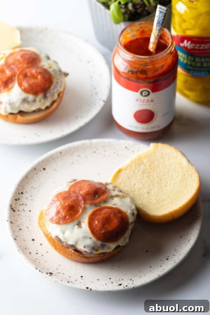 An open-face pizza burger proudly displayed on a white and black speckled plate, showcasing the bottom bun layered with sauce, a stacked burger patty, melted mozzarella, and pepperoni slices, inviting a closer look.