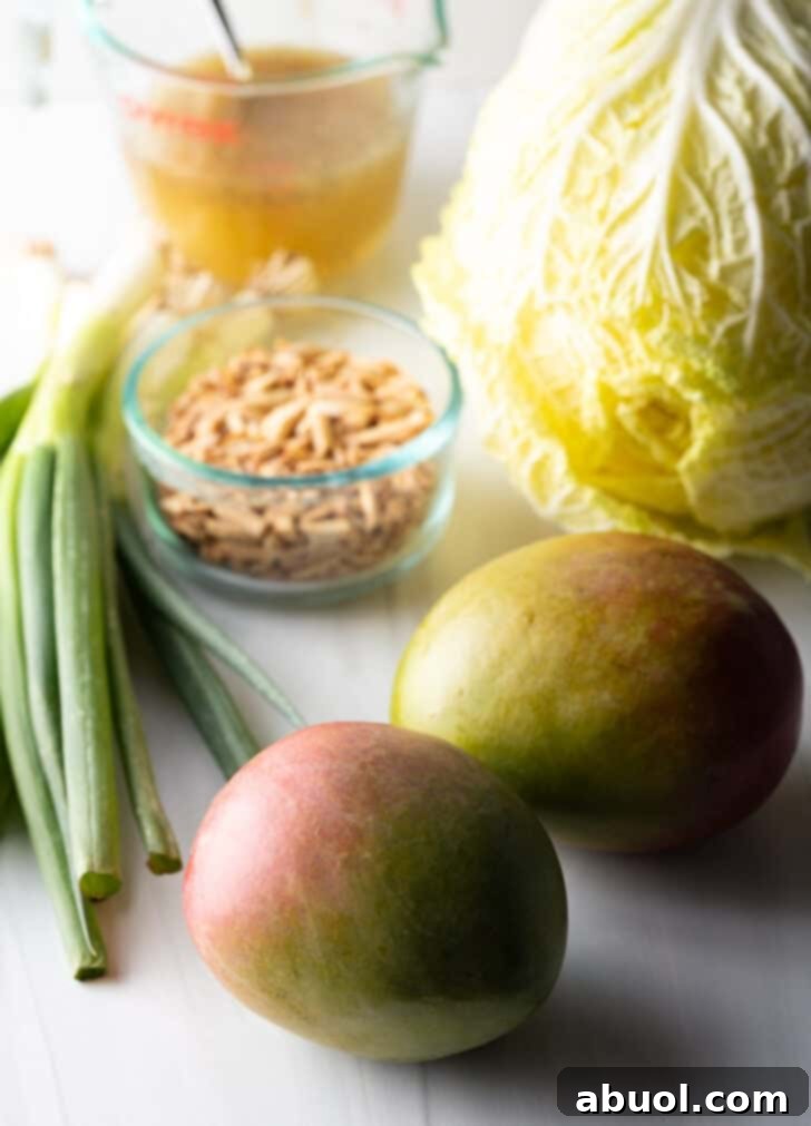 Napa cabbage and mango salad ingredients laid out: 2 whole ripe mangoes, a fresh bunch of green onions, a crisp head of napa cabbage, and a glass dish of golden toasted almonds.