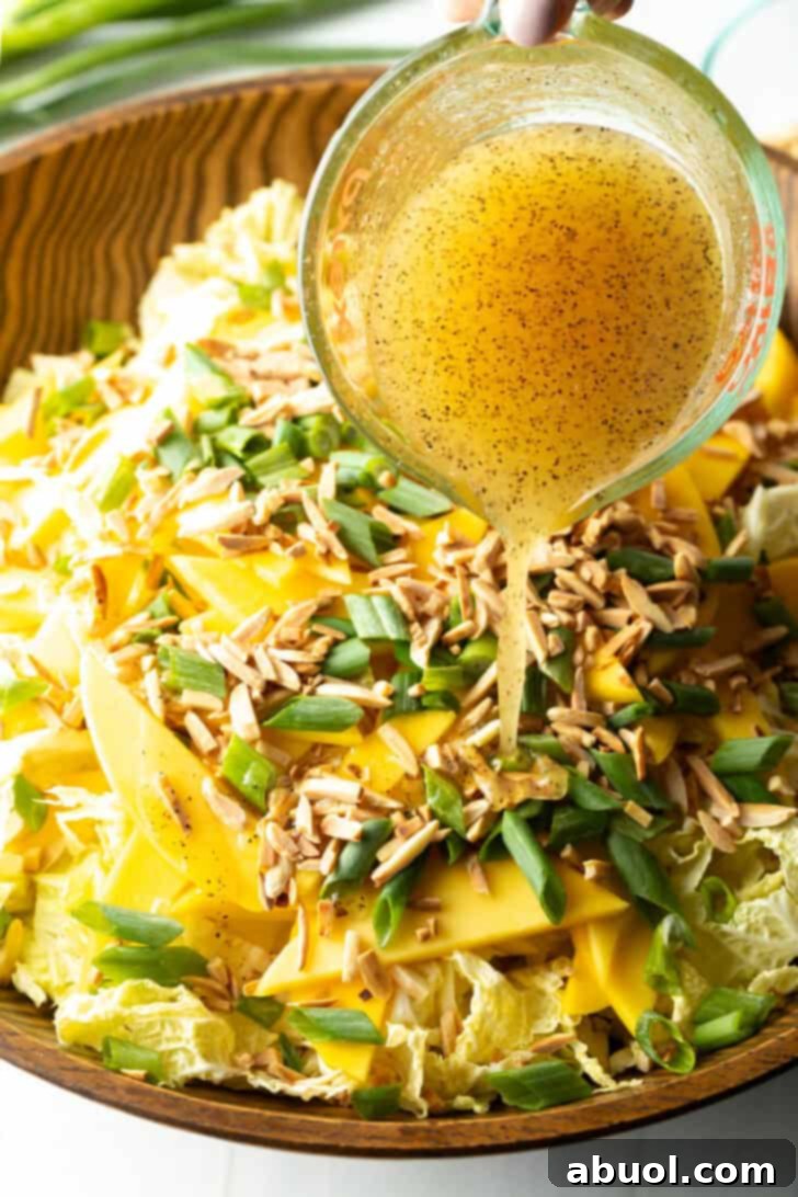 A skilled hand carefully pouring the glistening honey sesame vinaigrette from a glass measuring cup into a large bowl, enveloping the shredded cabbage, toasted almonds, green onion slices, and mango slices in its rich flavor.