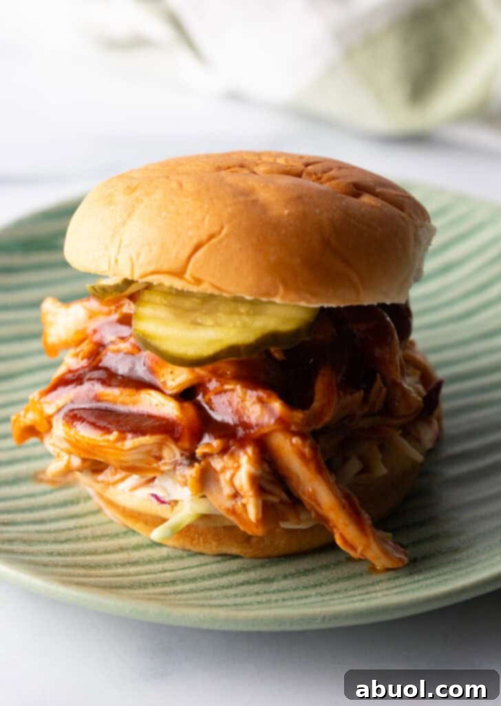 A mouth-watering crockpot BBQ chicken sandwich on a toasted hamburger bun, generously loaded with creamy coleslaw and tangy pickles.