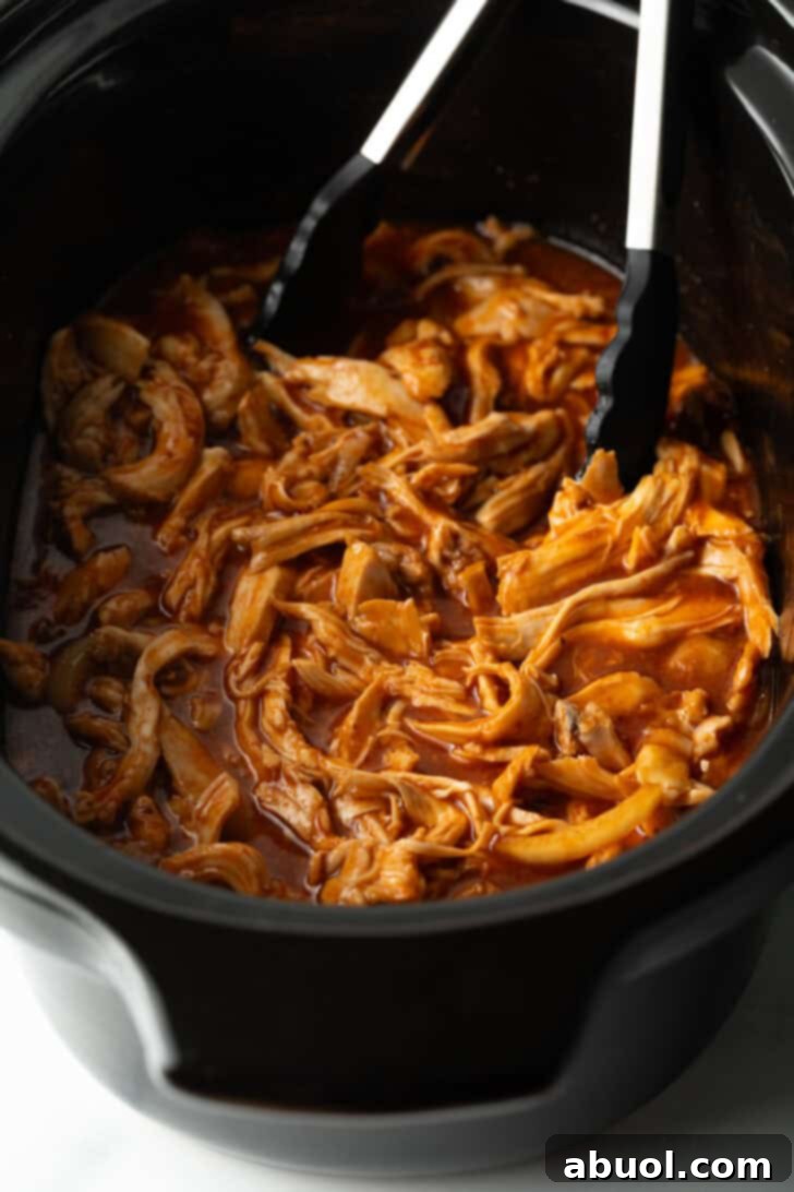 Tongs gently pulling apart tender pulled BBQ chicken in a black crockpot, showcasing its perfect shreddable texture, ready to be served.