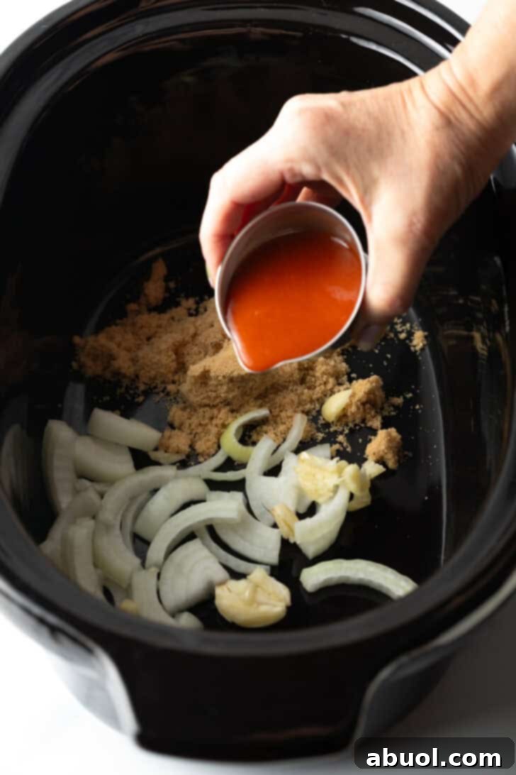 Pouring hot sauce from a small metal cup into the slow cooker, layering it over the brown sugar and sliced onions.