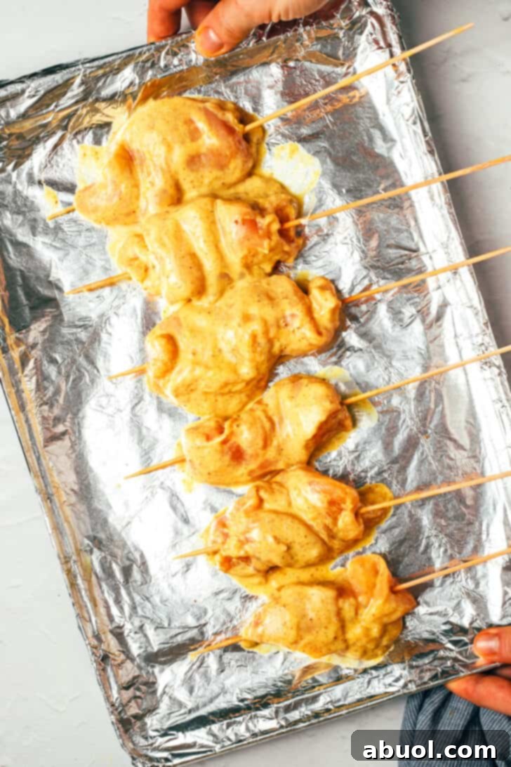 Hand holding a baking sheet covered with aluminum foil, holding 6 raw dipped chicken skewers.