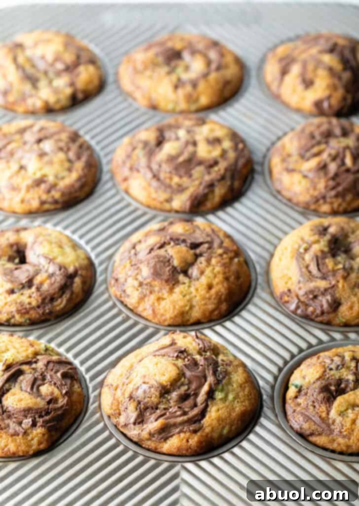 A close-up view of a dozen freshly baked zucchini and Nutella muffins still in their baking tin, showcasing their golden tops and enticing chocolate swirls.