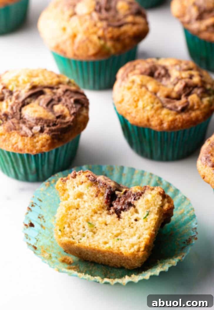 A stunning close-up of a dozen chocolate zucchini muffins with perfect Nutella swirls, presented in festive green cupcake wrappers. One muffin is thoughtfully sliced in half to showcase its fluffy, inviting interior.