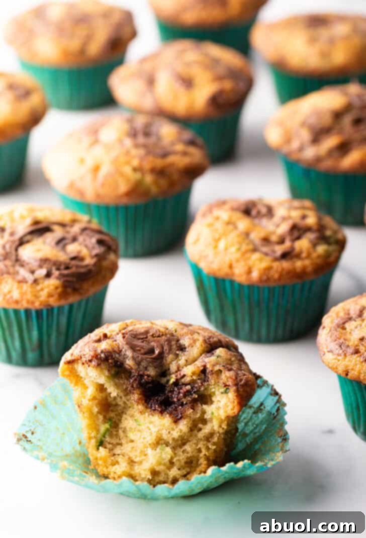 A dozen perfectly baked chocolate zucchini muffins with distinct Nutella swirls, nestled in green cupcake wrappers. One muffin is thoughtfully split to reveal its fluffy, moist interior.