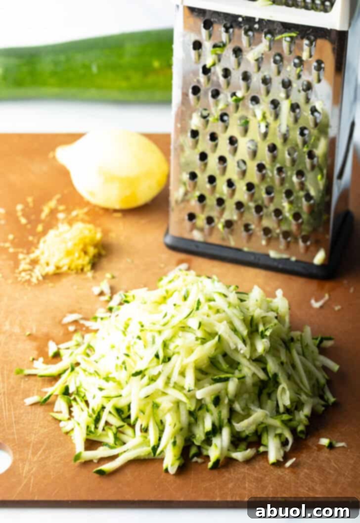 A box grater and a whole lemon alongside a generous pile of freshly shredded zucchini on a rustic wood cutting board, showcasing the preparation of key ingredients.