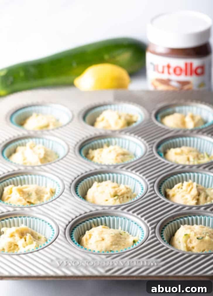 A 12-cup muffin tin meticulously lined with paper wrappers, each well evenly filled with chocolate zucchini muffin batter.