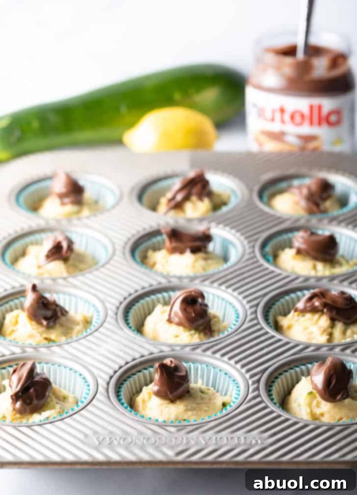 Raw chocolate zucchini muffin batter settled in a cupcake tin, each portion elegantly topped with a dollop of creamy Nutella, awaiting its swirl.
