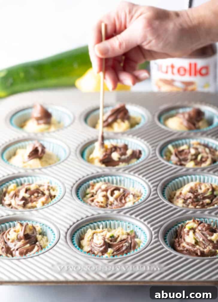 A hand expertly using a wooden skewer to artistically swirl Nutella into the unbaked zucchini muffin batter within a baking tin, creating beautiful marble patterns.